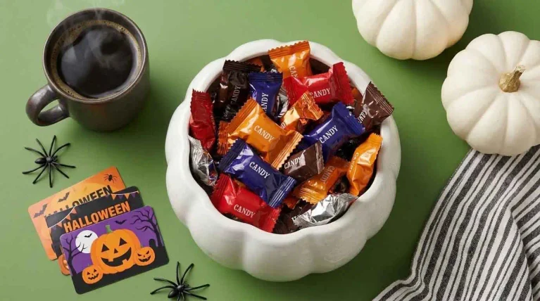 A white pumpkin-shaped bowl filled with colorful wrapped Halloween candy on a green table beside white pumpkins and Halloween decorations.