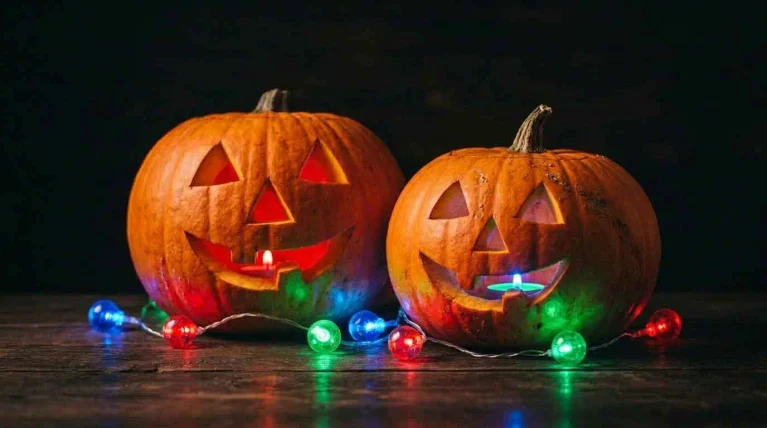 Two glowing carved Halloween pumpkins beside colorful string lights on a dark wooden surface.